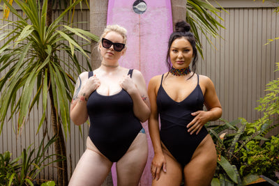 Models wearing SWMR The Dunk one piece and the Dip one piece swimsuits in black standing with a surfboard